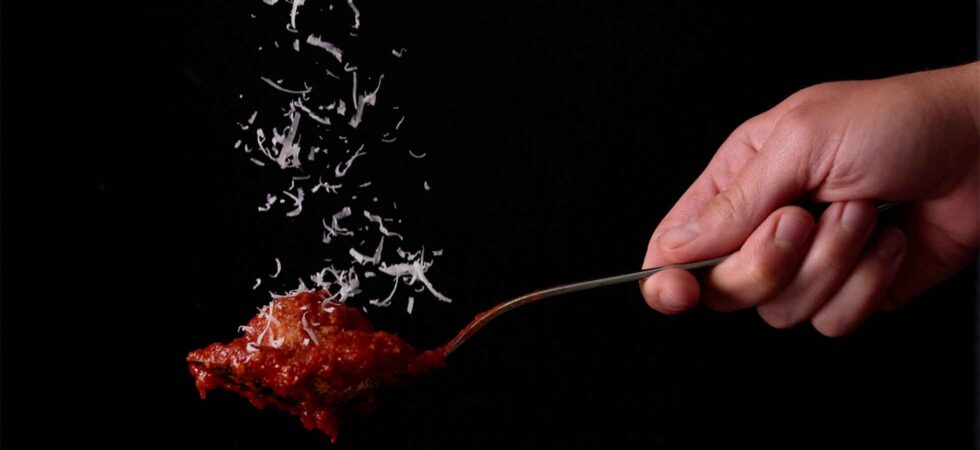 Italian meatball, covered in sauce, on a spoon. Parmesan is sprinkling down from the top of the image to rest on the meatball.