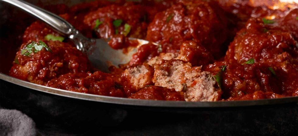 Italian meatballs and tomato sauce in a sauté pan. One meatball is cut in half so you can see it;s juicy center.