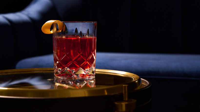 Negroni in a glass with a clear ice cube and orange peel garnish sitting on a side table made of glass and brass. A blue velvet couch is in the background.