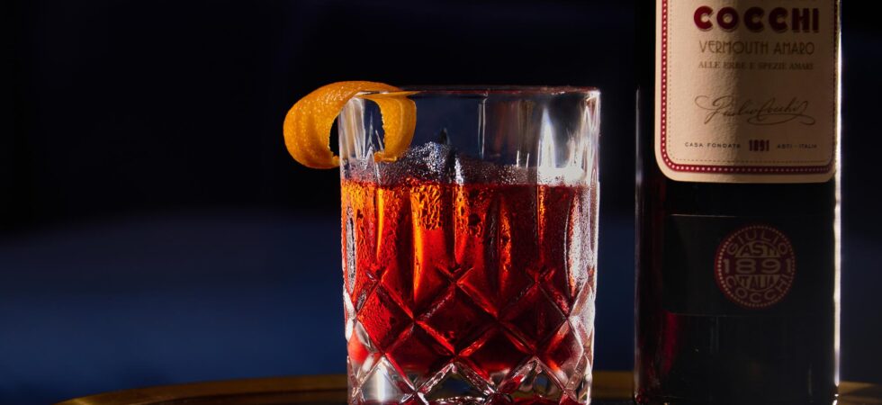 Night Cap Negroni in a glass with a clear ice cube and orange peel garnish. A bottle of Cocchi Dopo Teatro Vermouth amaro next to it.