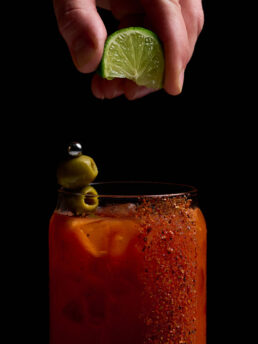 Squeezing a lime wedge into a Bloody Mary. The glass has tajin running down the side and an olive skewer garnish.