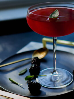 Mezcal blackberry shrub cocktail in a coupe glass and dappled light, with a sage leaf garnish.