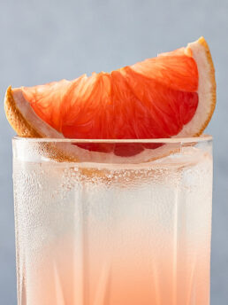 Up-close, grapefruit wedge place on top of a paloma cocktail in a highball glass.