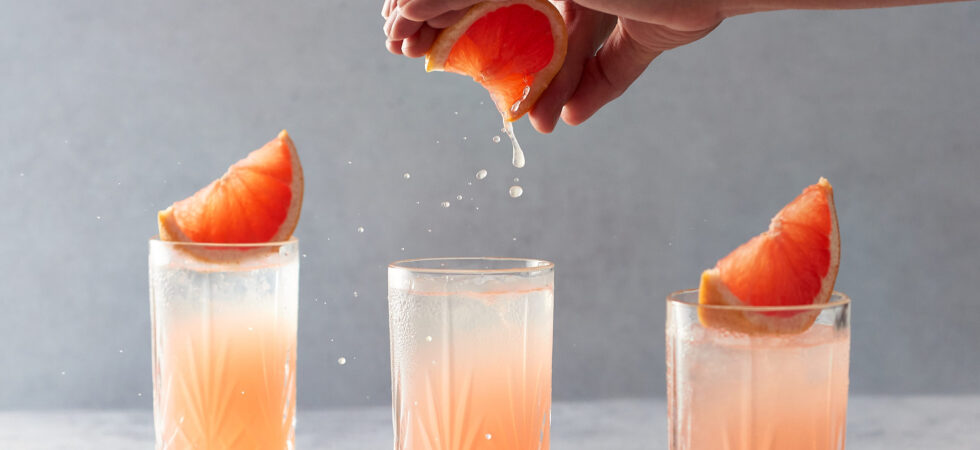 Squeezing a grapefruit over a paloma cocktail in a highball glass