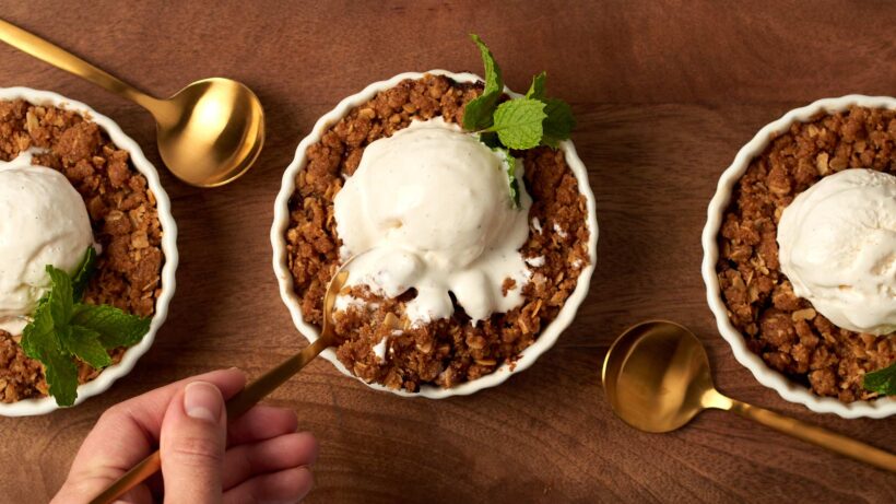 The first scoop from a nectarine crisp. Three nectarine crisps in ramekins, in a row, all topped with a scoop of ice cream and a sprig of mint. The scoop is being taken from the center ramekin.