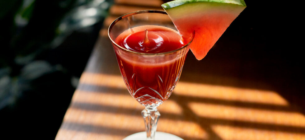 A closeup of a Watermelon-Tamarind Cognac Cocktail with a single droplet splashing up out of a Nick & Nora glass.