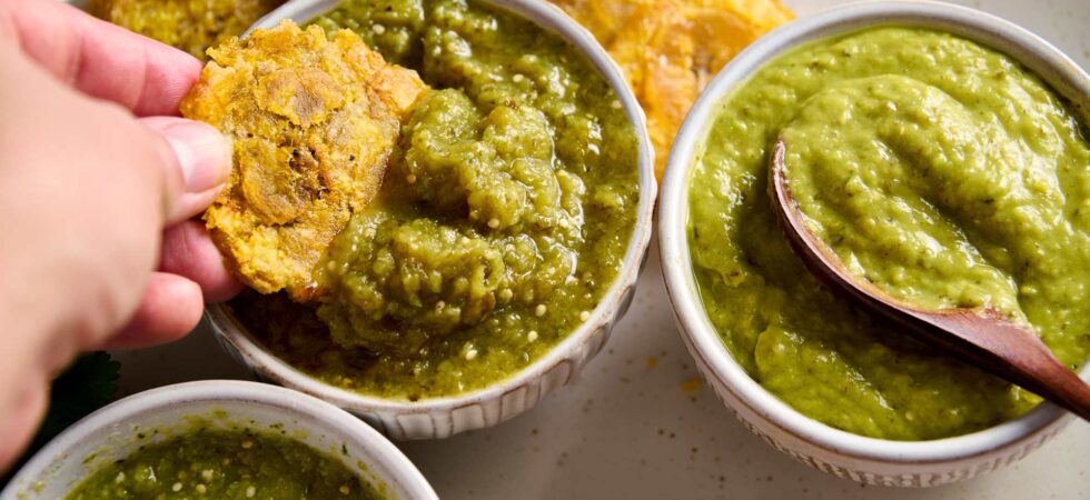 Three different variations of salsa verde, in dip bowls, with a hand dipping a tostone into one of them.