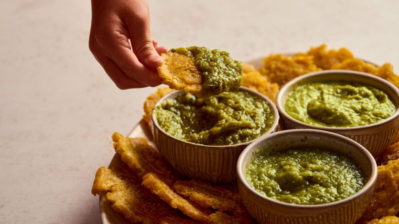 Three different tomatillo salsas, in dip bowls, with a hand dipping a tostone into one of them.