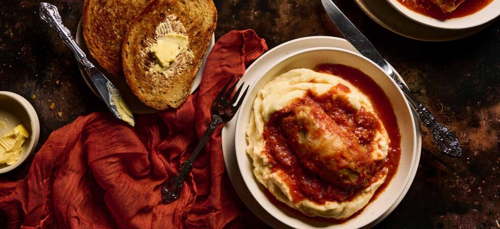 A stuffed cabbage roll plated on a bed of mashed potatoes with toasted, buttered rye bread.