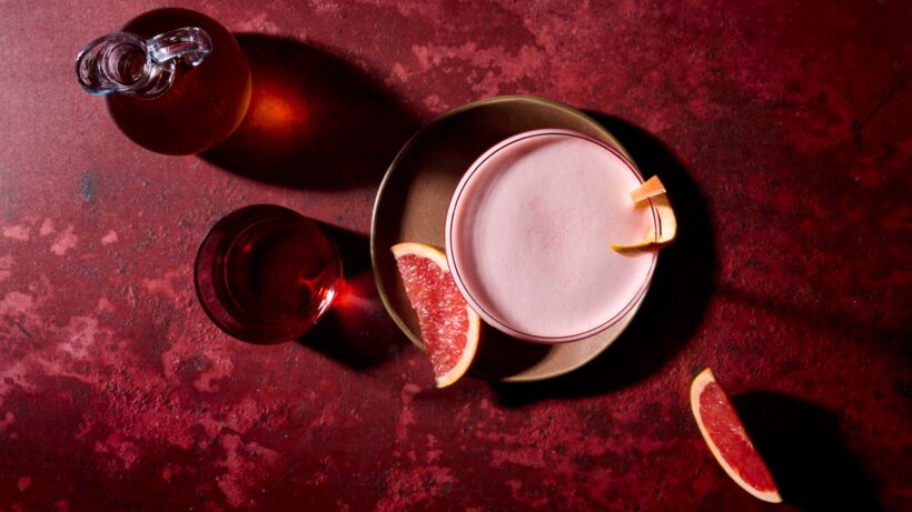 A top down tablescape of a grapefruit-champagne negroni sour in coupe glass with a grapefruit peel garnish. Sliced grapefruit wedges, a glass of Campari, and a bottle of Champagne syrup surround the cocktail.