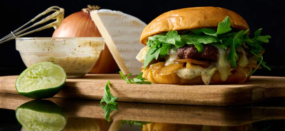 gourmet griddled onion and brie burger on a wooden cutting board surrounded by ingredients.