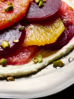 Closeup of a marinated beet and citrus salad, with sliced beets and citrus, on a bed of whipped goat cheese and topped with chopped pistachios.