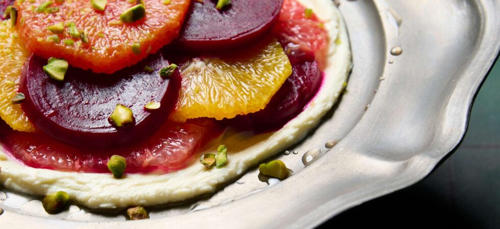 Closeup of a marinated beet and citrus salad, with sliced beets and citrus, on a bed of whipped goat cheese and topped with chopped pistachios.